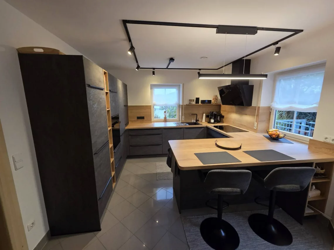 Gesamtansicht einer modernen Wohnküche mit robusten Stone-Basalt-Fronten, Arbeitsplatte in Holzstruktur, großem Sitzbereich, hochgebautem Backofen und großzügigen Stauraumlösungen.